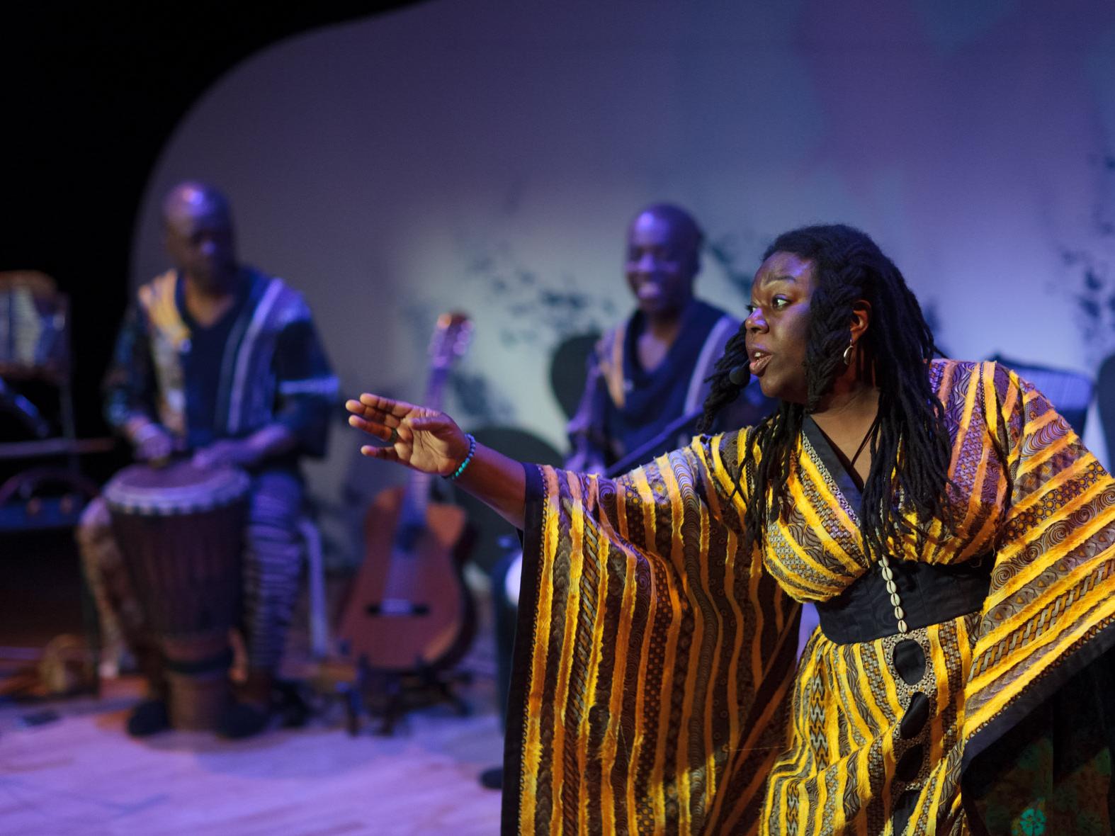 storyteller Jan Blake wearing a gold and yellow costume stretches out her hand, musicians Raymond & Kouame Sereba can be seen in the background