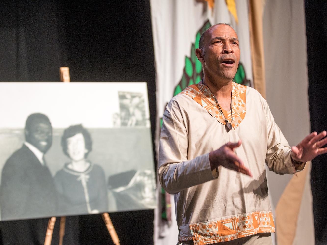 Phil Okwedy is standing on a stage looking out to the audience, with his arms raised up in front of him, and is stood in front a photograph of his parents. This is the only photo he has of his Nigerian Father and his Welsh mother before his father moved back to Nigeria.