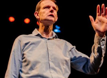 a white man with short brown hair, wearing a blue shirt and gesturing with his hand