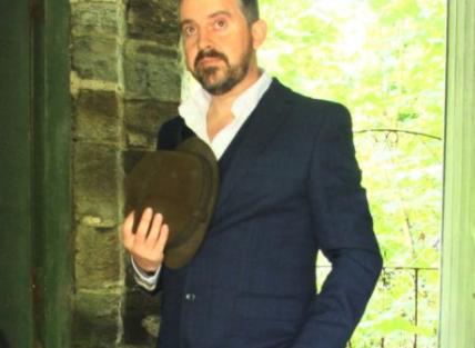 a white man with short dark hair and beard, wearing a suit and holding a hat