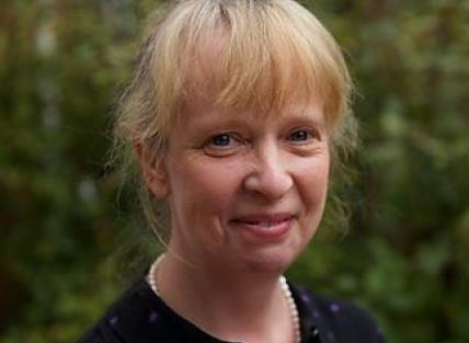 a white woman with blond hair tied back, wearing a blue dress and pearl necklace